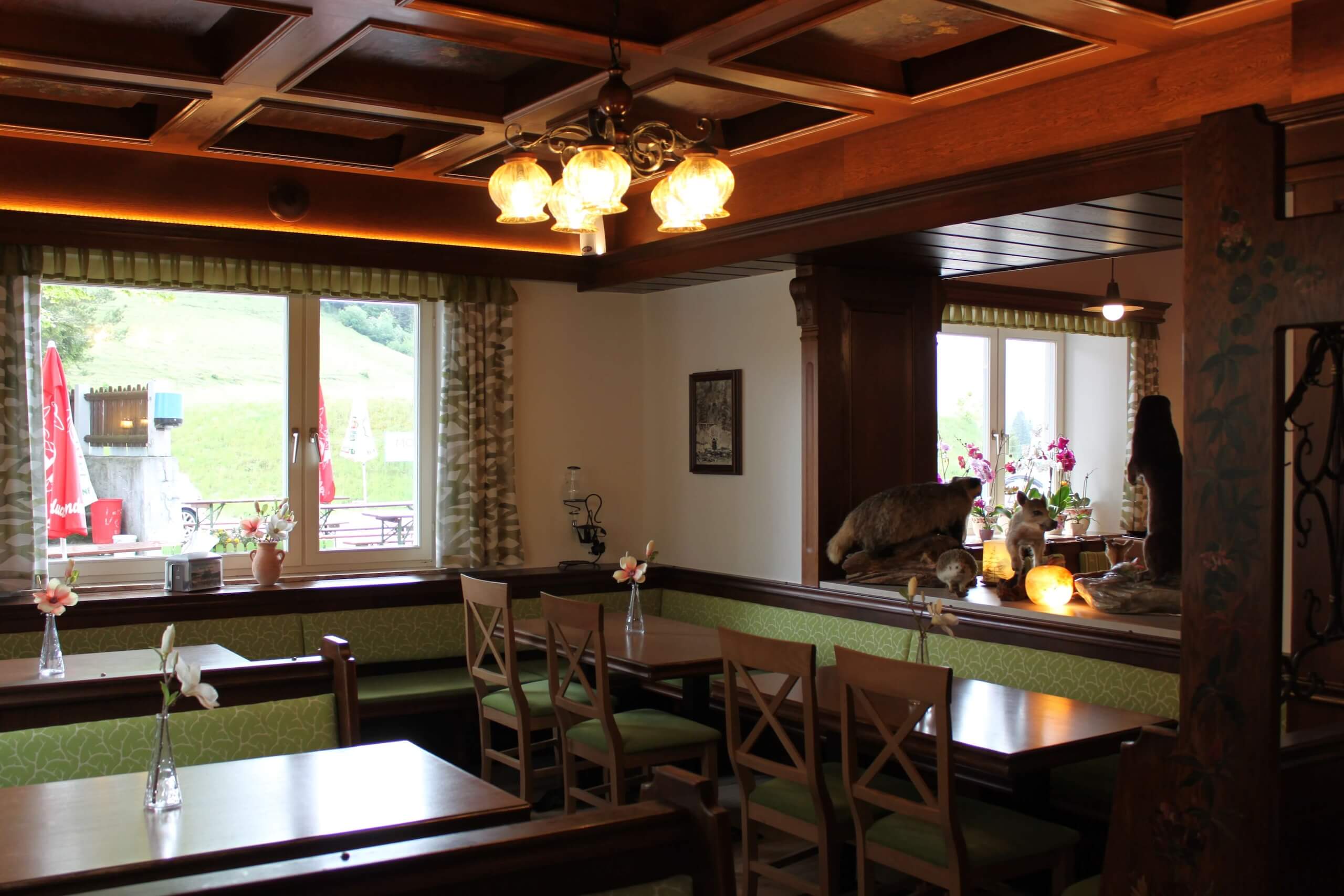Gemütliche Gaststube mit Holzmöbeln, dekoriert mit Blumen und einem Blick auf die malerische Landschaft durch große Fenster.
