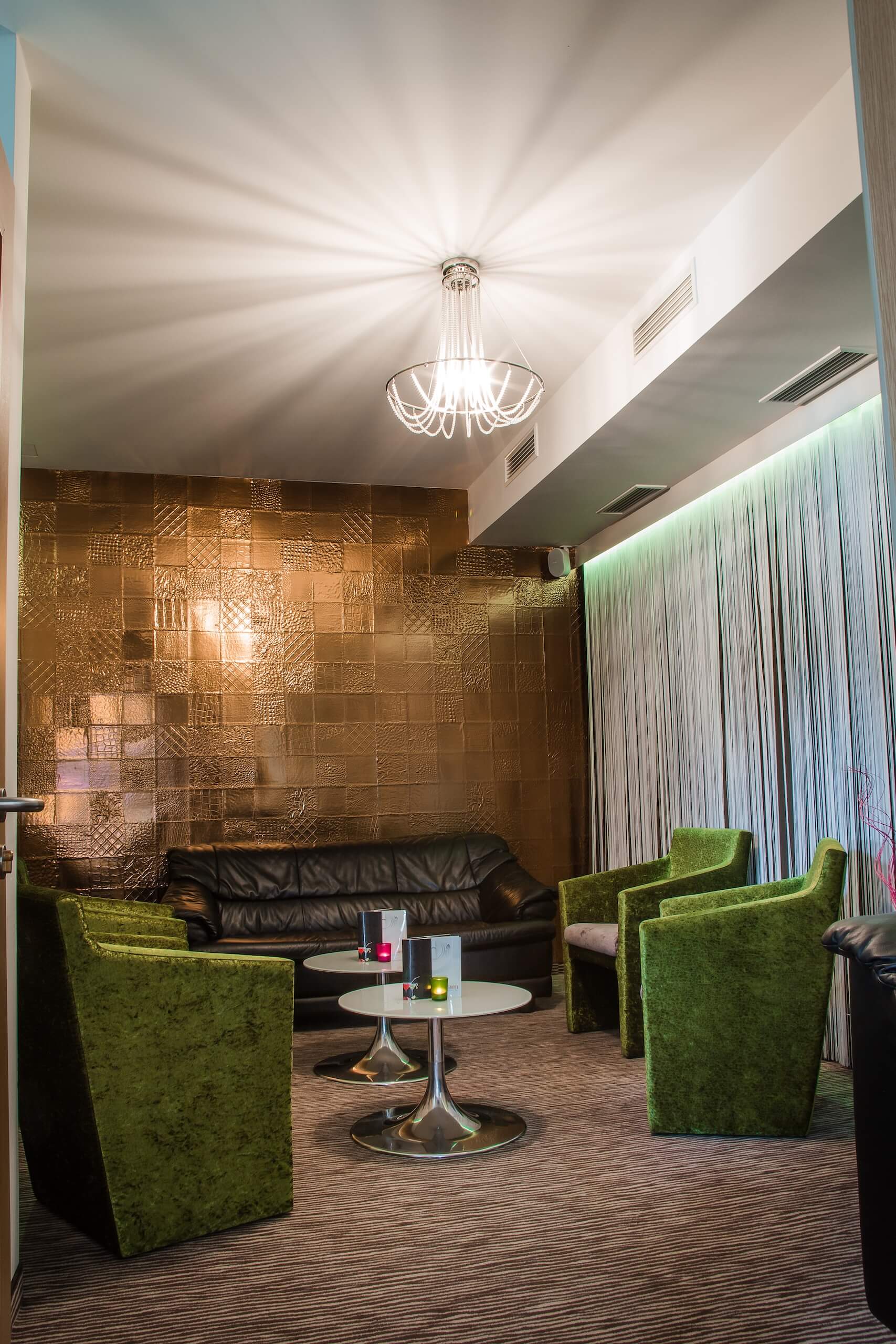 Elegantes Wohnzimmer mit grünen Sesseln, Ledersofa und strahlendem Kronleuchter vor goldener Akzentwand.