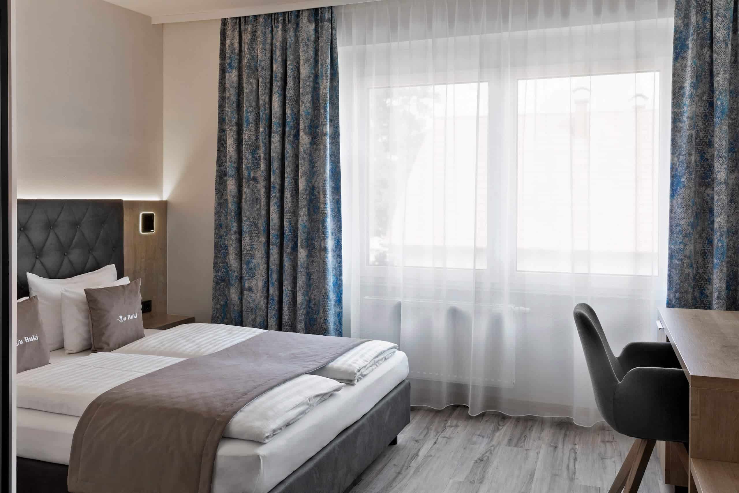 Modern eingerichtetes Hotelzimmer mit Doppelbett, Schreibtisch und großen Fenstern mit blau-gemusterten Vorhängen.