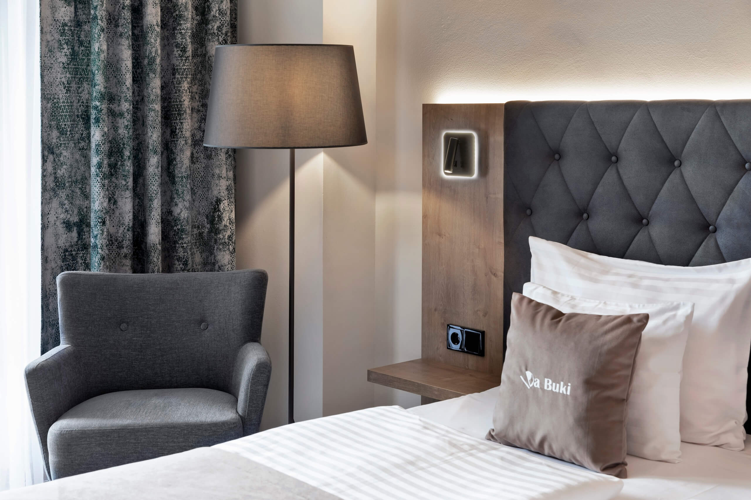 Elegantes Hotelzimmer mit Komfortbett, moderner Lampe und stilvollem Sessel neben einer großen Fensterfront.