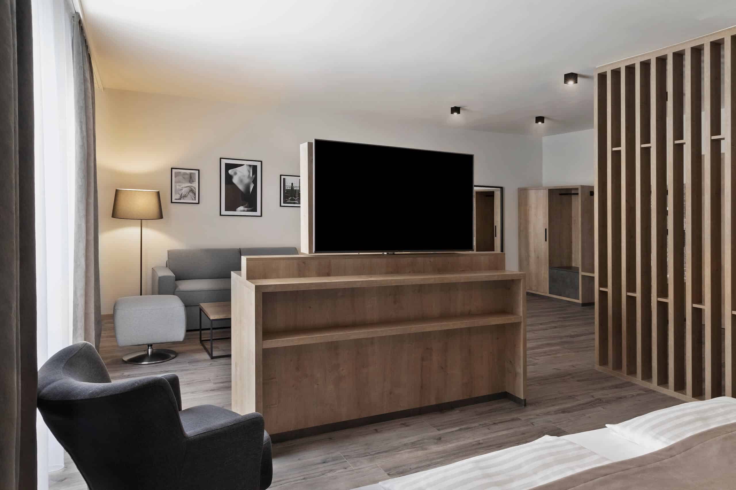 Modernes Hotelzimmer mit Holzdekor, Sofa, Sessel, Flachbild-TV, Kunstwerken und Stehlampe.