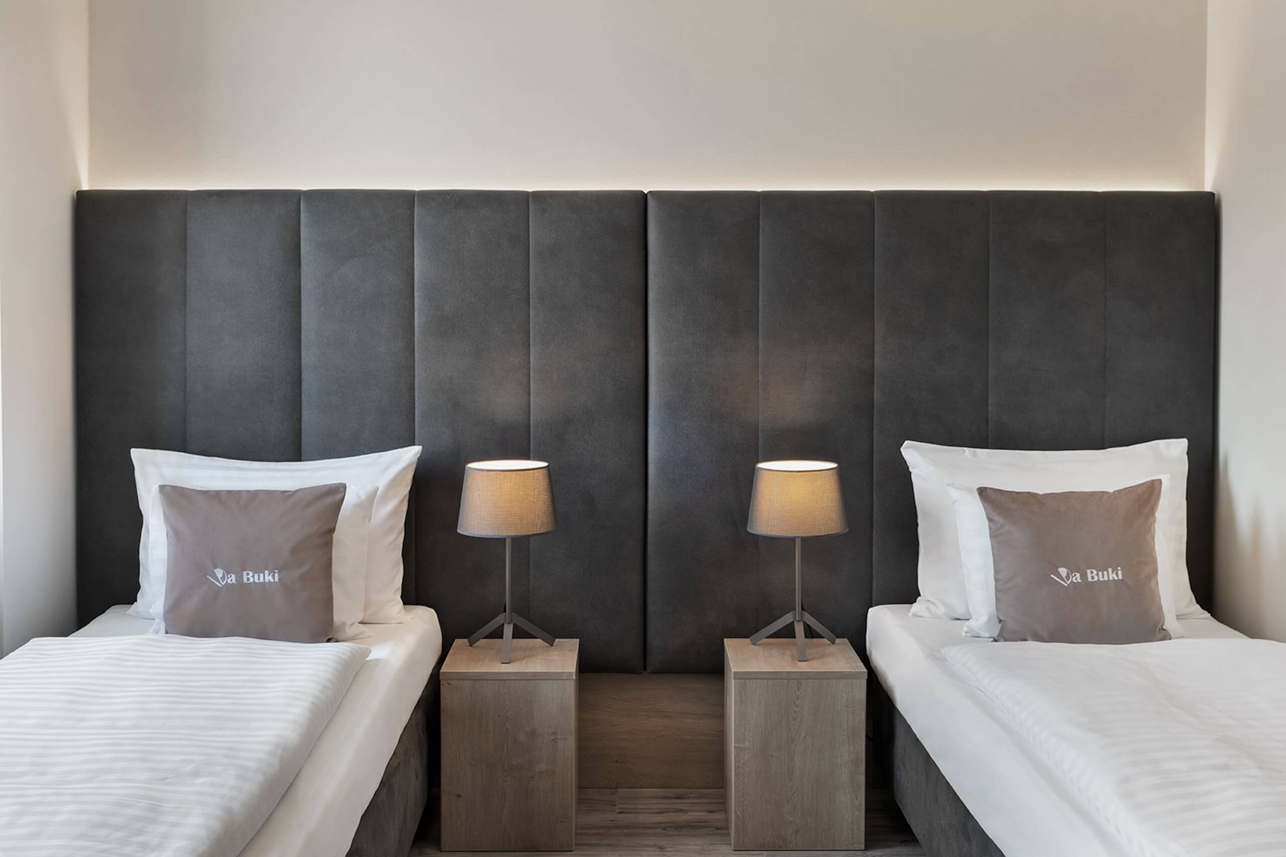 Moderner Hotelraum mit zwei Einzelbetten, dunklem Kopfteil und Nachttischlampen aus Holz für eine gemütliche Atmosphäre.