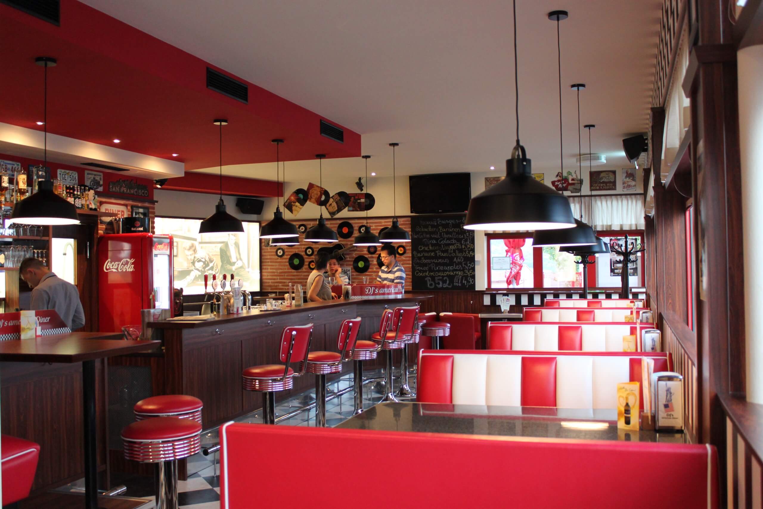 Retro 50er-Jahre Diner mit roten Sitzbänken, Barhockern und Coca-Cola-Kühlschrank; gemütliche Atmosphäre.