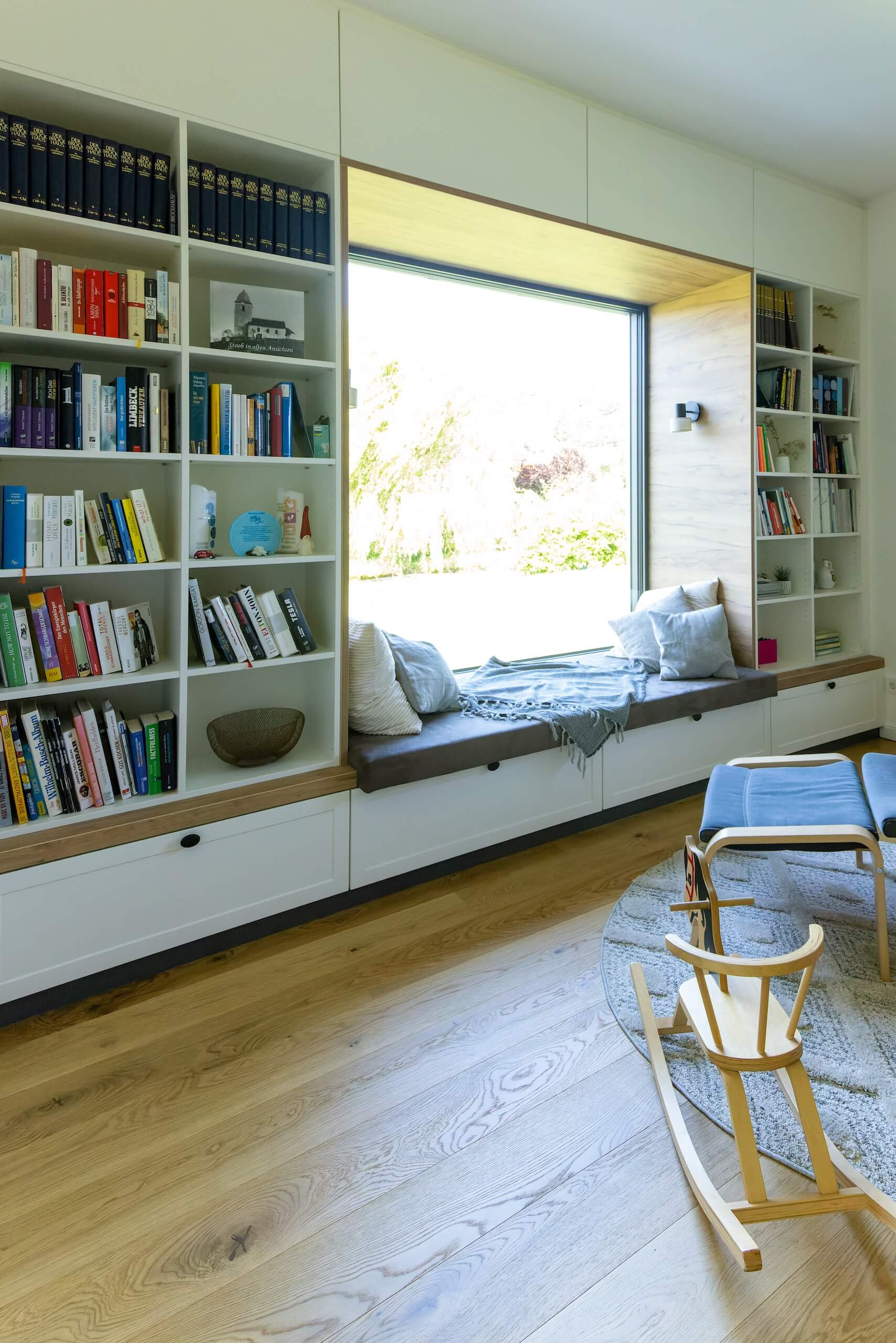 Gemütliche Leseecke mit Bücherregal, großem Fenster und Sitzbank mit Kissen für entspannende Lesestunden.