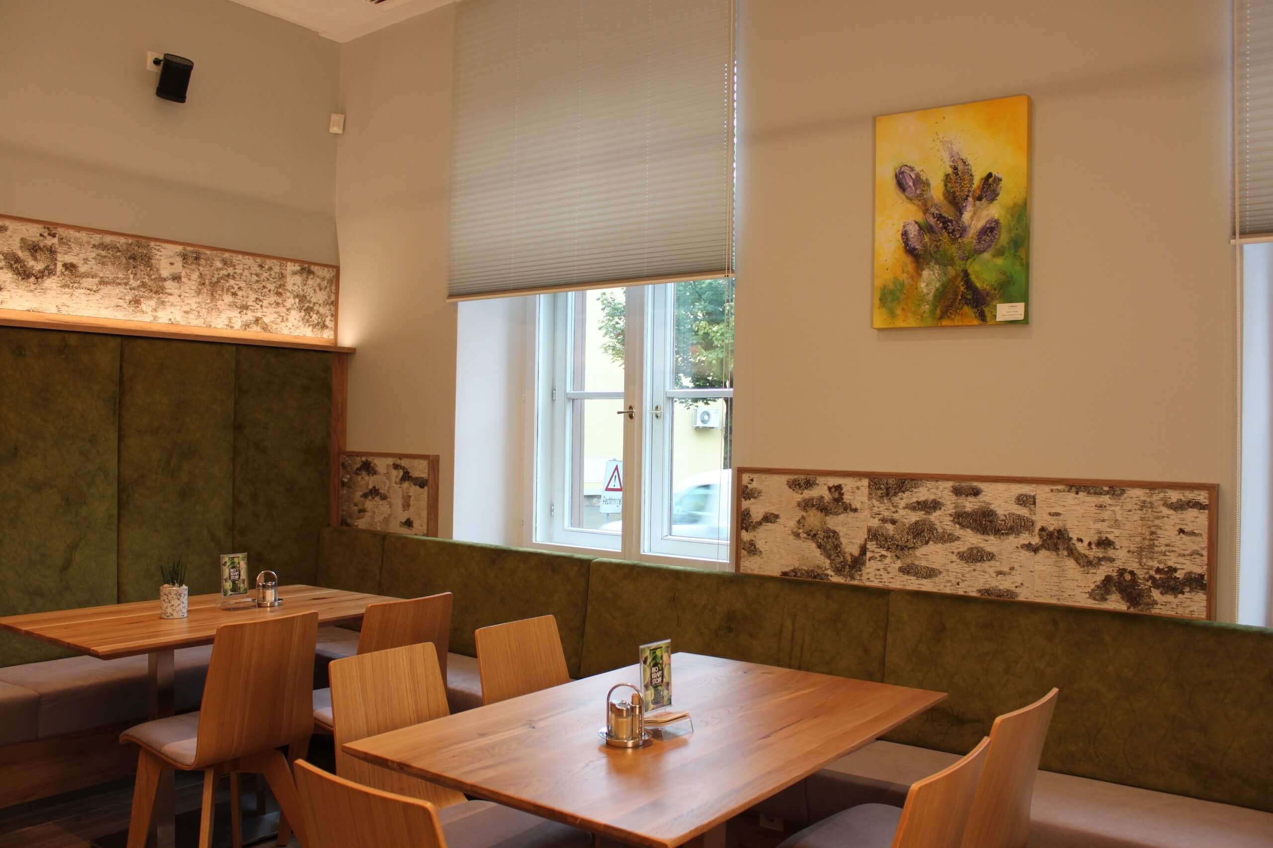 Moderne Café-Innenansicht mit Holztischen, grünen Sitzbänken und einem hellen Fenster für gemütliches Ambiente.