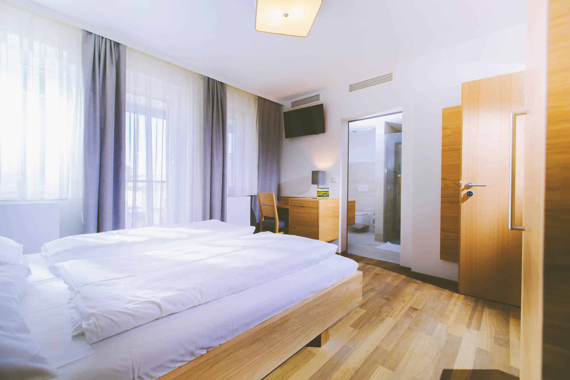 Helles Hotelzimmer mit großen Fenstern, Doppelbett, Holzmöbeln und Zugang zu einem modernen Badezimmer.