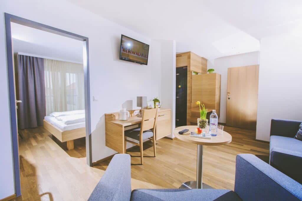 Modernes Hotelzimmer mit hellem Holzdesign, Schreibtisch, Fernseher und gemütlichem Sitzbereich im Wohnstil.