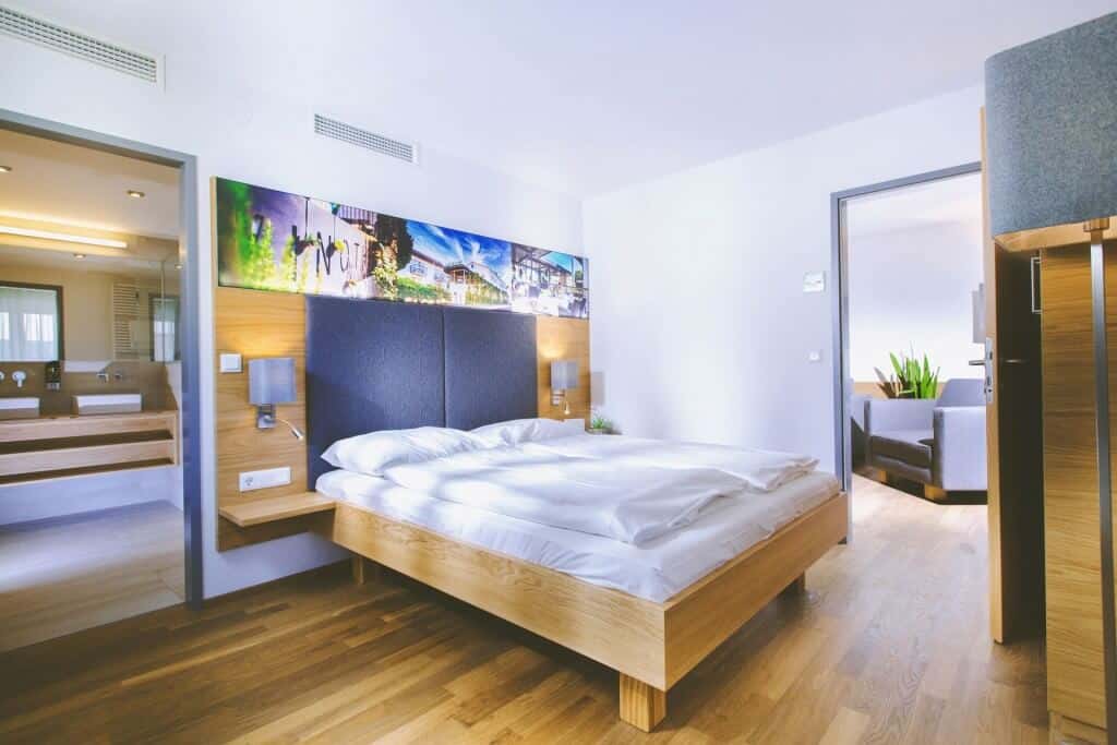 Modernes Hotelzimmer mit Doppelbett, hellem Holzfußboden und Blick auf Badezimmer und Wohnzimmer im Hintergrund.