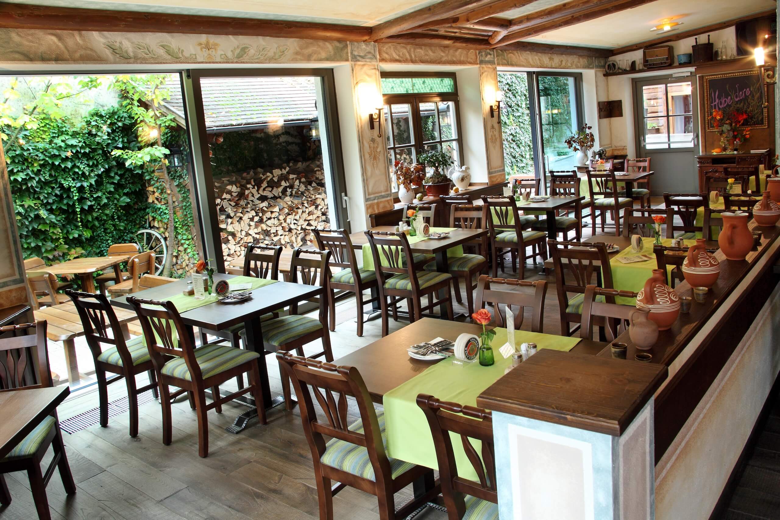 Gemütliches Restaurant mit Holzdekor, grünen Tischläufern und Gartenblick durch große Fenster im Hintergrund.