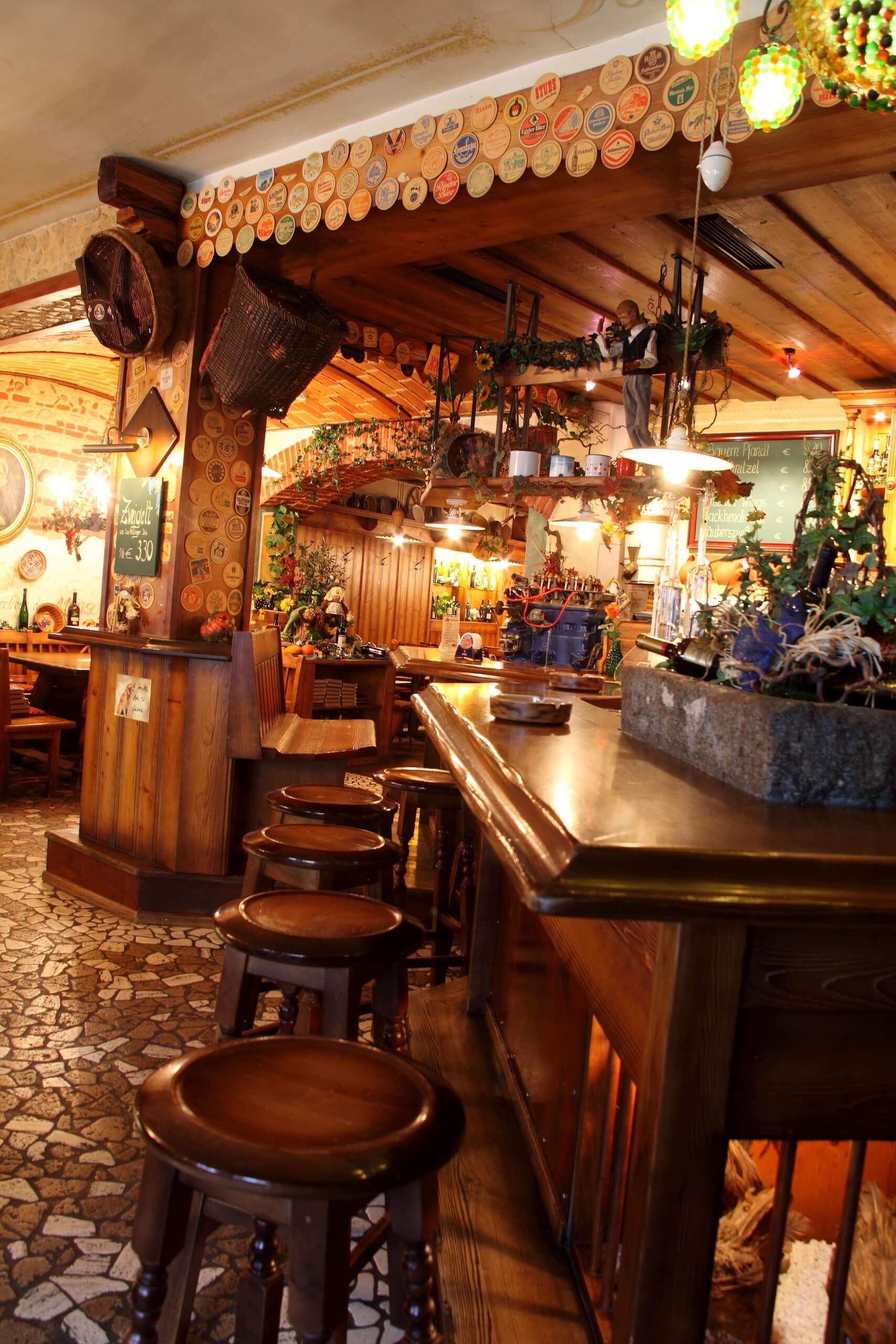 Gemütliche Kneipe mit Holztresen und Barhockern in rustikalem Stil, dekoriert mit Bierdeckeln an der Decke.