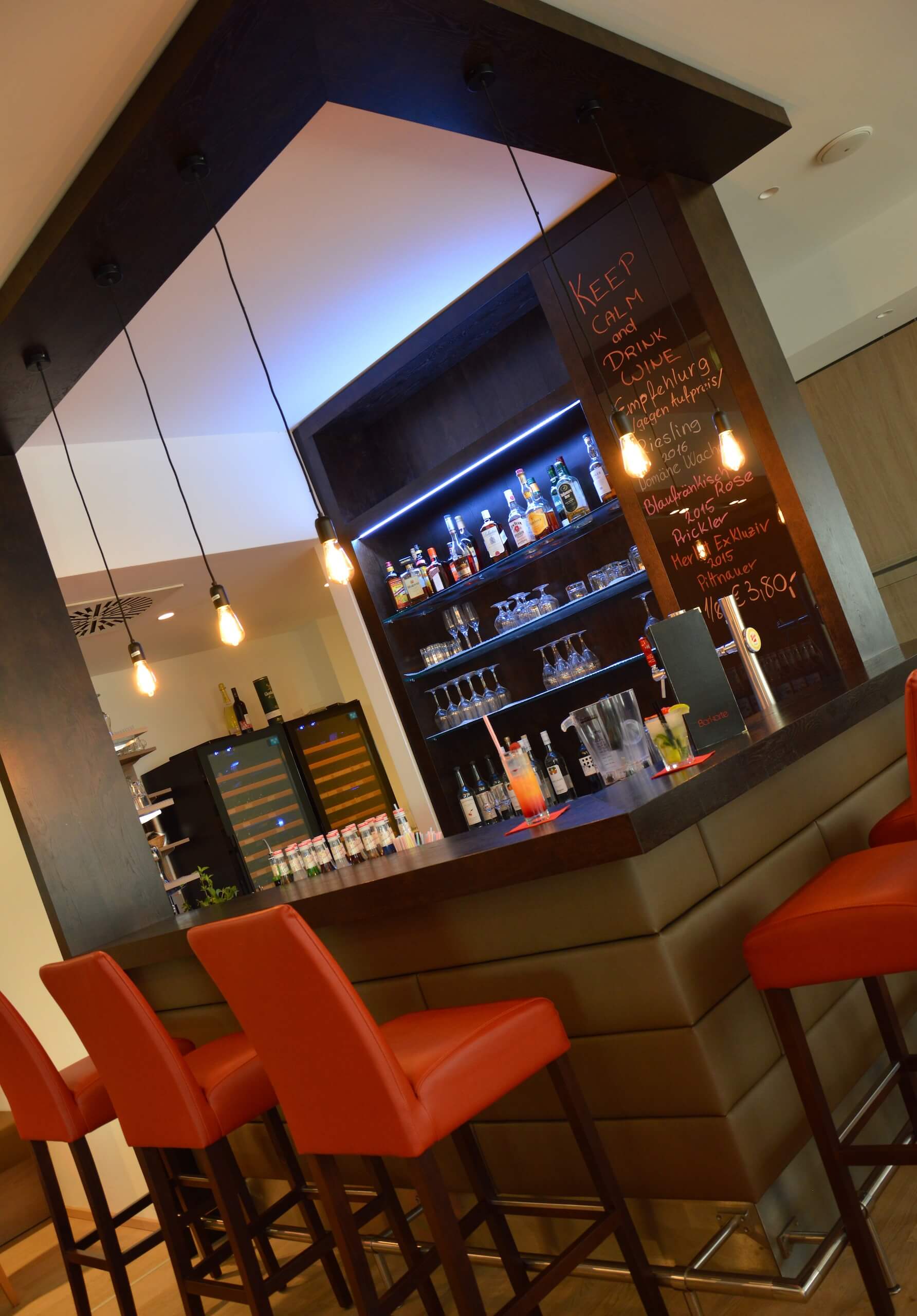Modern gestaltete Bar mit roten Hockern, beleuchteten Regalen und dekorativer Getränketafel für ein stilvolles Ambiente.