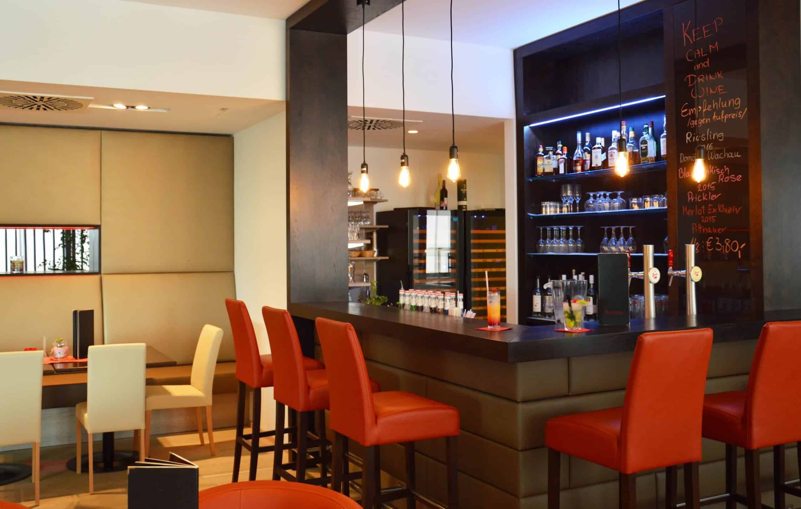Moderne Hotelbar mit roten Hockern und beleuchtetem Flaschenregal im Hintergrund. Gemütliches Ambiente für Drinks.