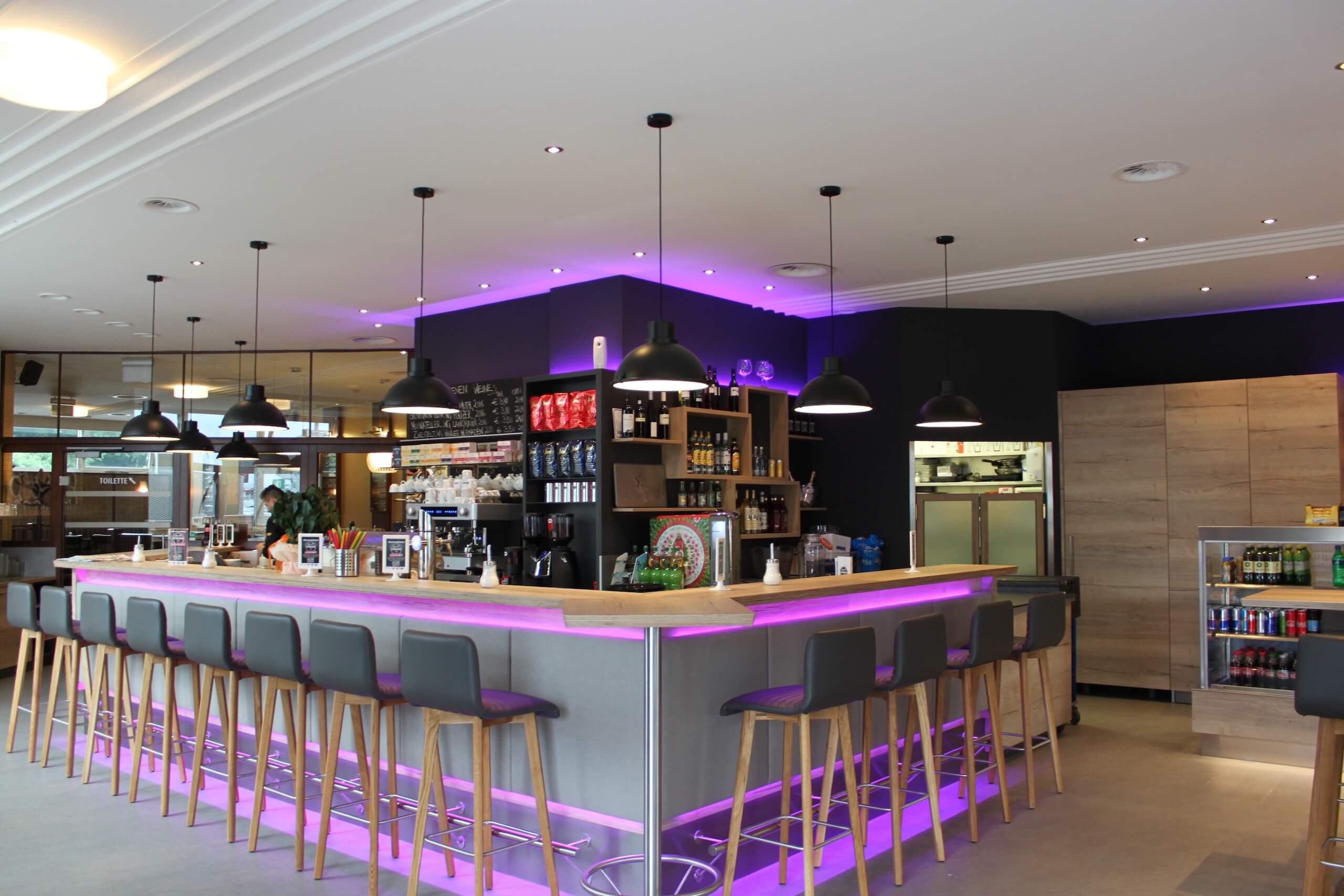 Moderne Cafébar mit lila Beleuchtung, Barhockern und Regalen voller Getränke und Kaffeezubehör.