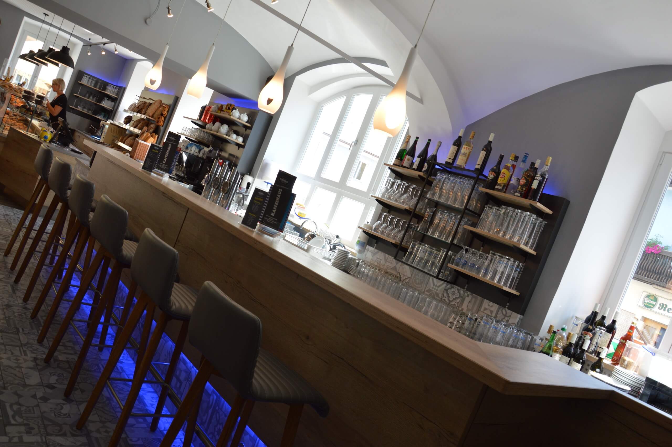 Moderne Café-Bar mit hölzerner Theke, Barhockern und eleganter Beleuchtung vor großen Fenstern.
