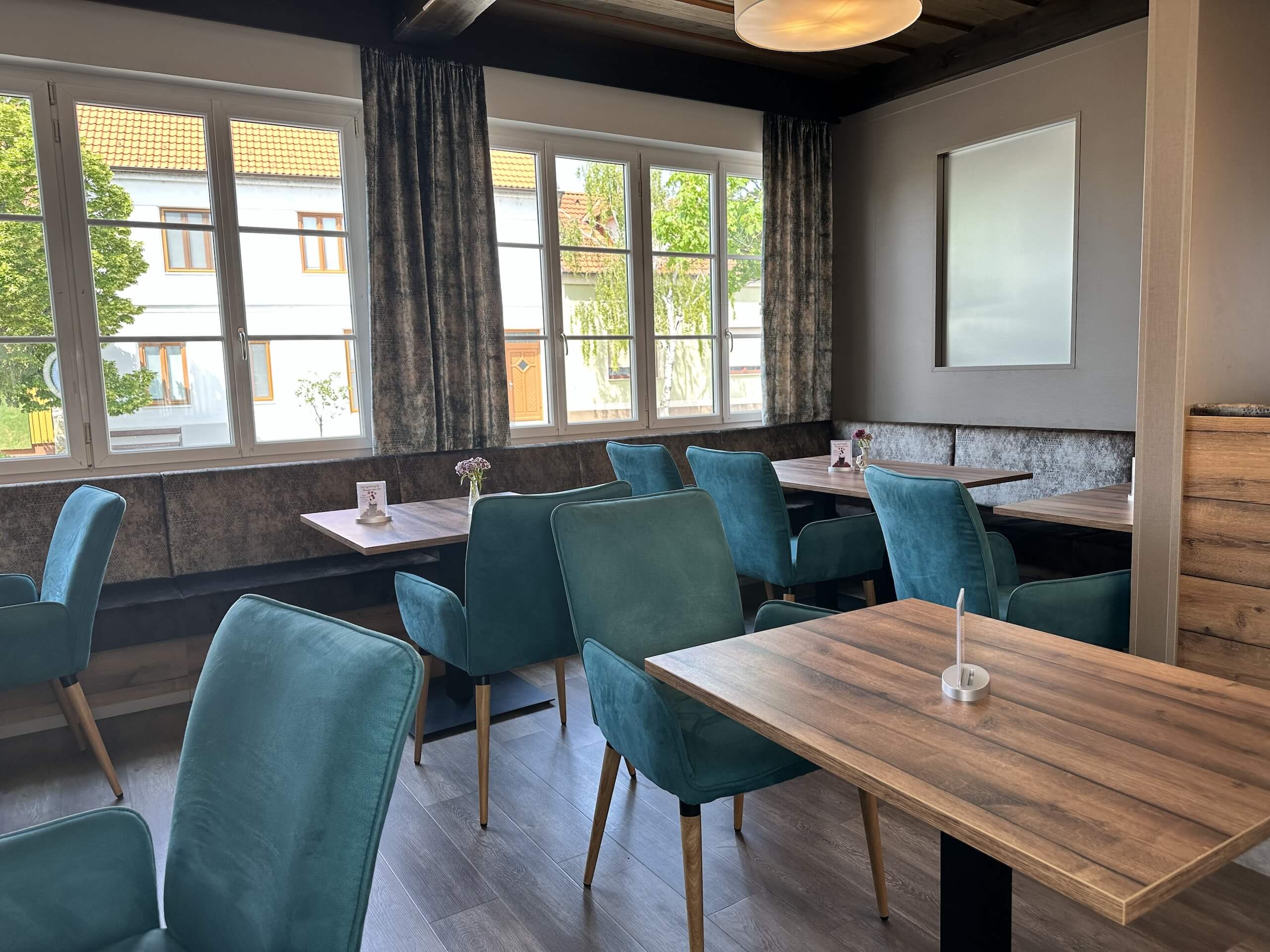 Moderne Caféeinrichtung mit türkisfarbenen Stühlen und Holztischen, großes Fenster mit Blick nach draußen.