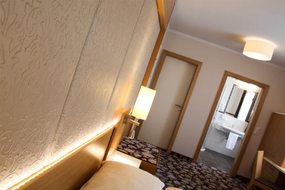 Modernes Hotelzimmer mit beleuchteter Tapete, Bettlampe und Zugang zum Badezimmer, ideal für eine entspannte Übernachtung.