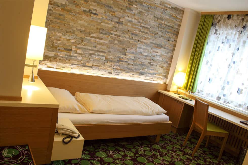 Modernes Hotelzimmer mit Steinwand, Einzelbett, Schreibtisch und Fenster mit grünen Vorhängen und Tageslicht.