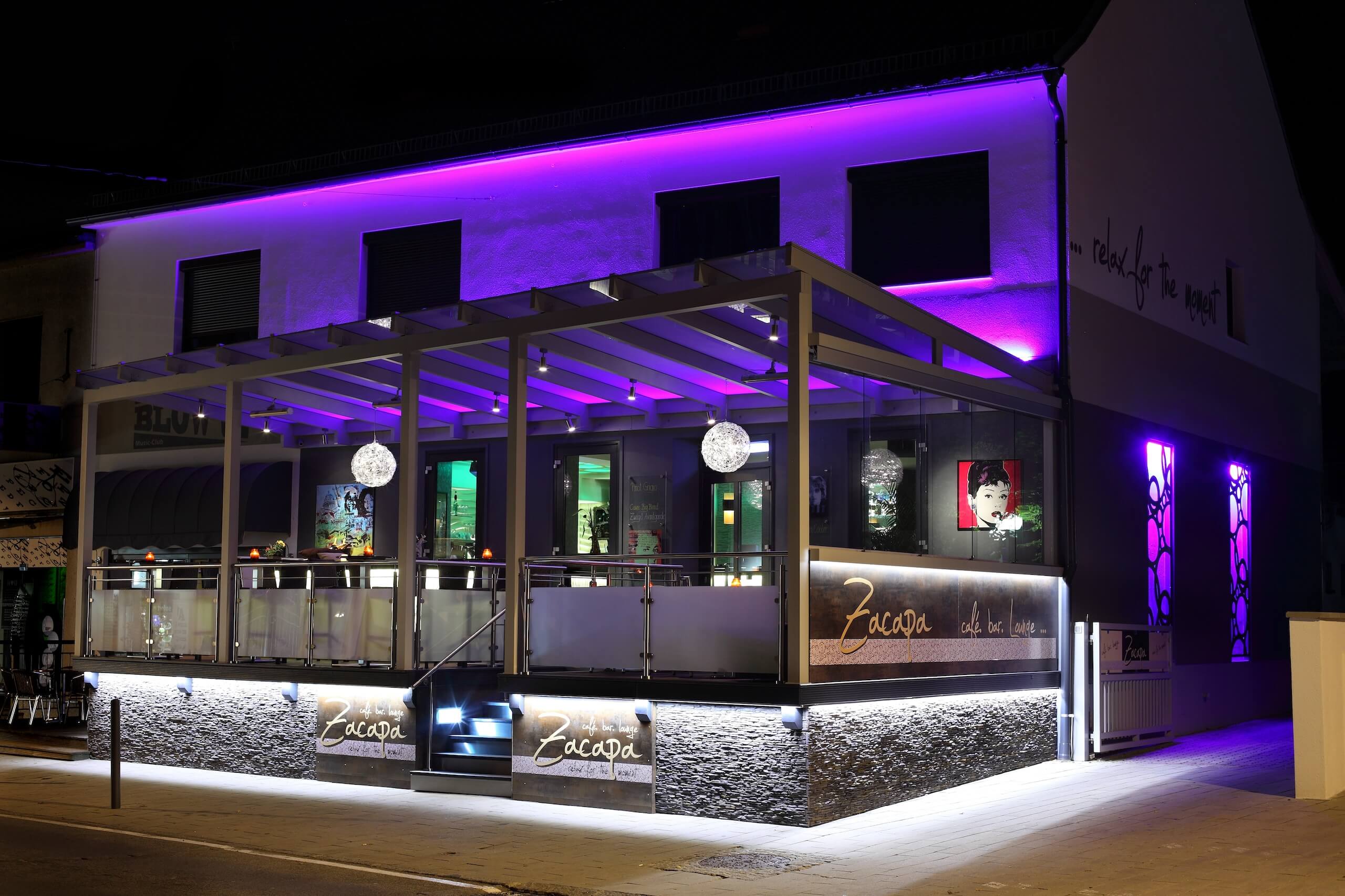 Beleuchtetes Café bei Nacht mit lila Akzentlicht und eleganter Außenterrasse.