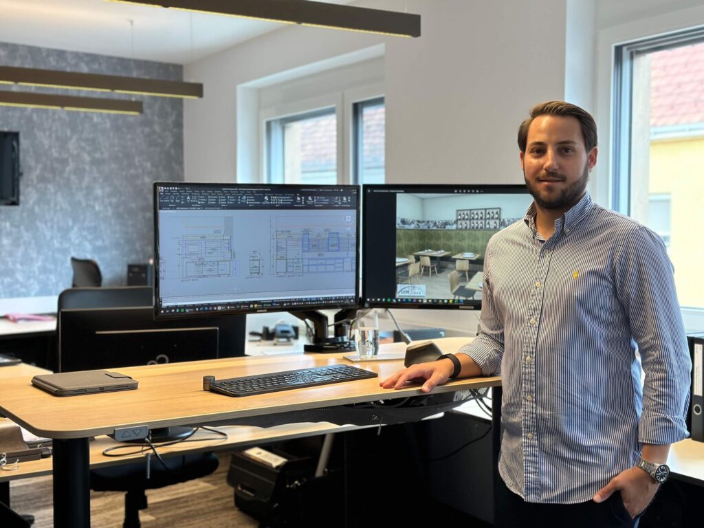 Mann vor zwei Monitoren im Büro mit Architekturzeichnungen, lässig gekleidet, modernes Arbeitsumfeld.